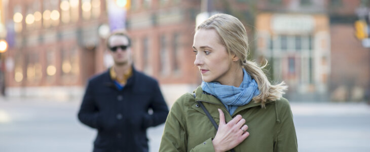 Scared young woman with blonde hair looking back over her shoulder at a stranger in a black trench coat that is following behind her downtown in an urban city.